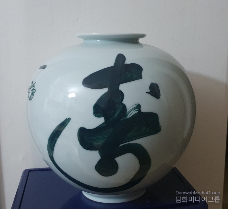 「壽」(수) – 일붕 서경보 법왕 친필 휘호 백자 항아리, 이 작품은 초대 법왕 일붕 서경보一鵬 徐京保)스님께서 친히 휘호하신 「壽」(수, ‘목숨 수’) 자를 백자 항아리에 담은 귀중한 예술품이다. '壽'는 장수, 생명, 복덕의 근원을 상징하며, 불가佛家에서는 중생의 안녕과 평안을 기원하는 자비의 상징어로 자주 사용되어 왔다. 강건하고 유려한 필획으로 휘호된 이 글씨는 단순한 '장수'의 의미를 넘어, 생명을 향한 경외, 불변의 진리를 향한 염원을 품고 있다. 백자의 순백 위에 검푸르게 펼쳐진 붓결은 일붕 스님의 정신성과 예술성이 응축된 선묵禪墨의 정수라 할 수 있다. 본 항아리는 현재 담화총사 소장으로, 수행과 기도, 예술과 정신의 통합적 상징물로 여겨지며, 스님의 자비 사상과 호흡을 함께 느낄 수 있는 성물로서의 가치를 지닌다.